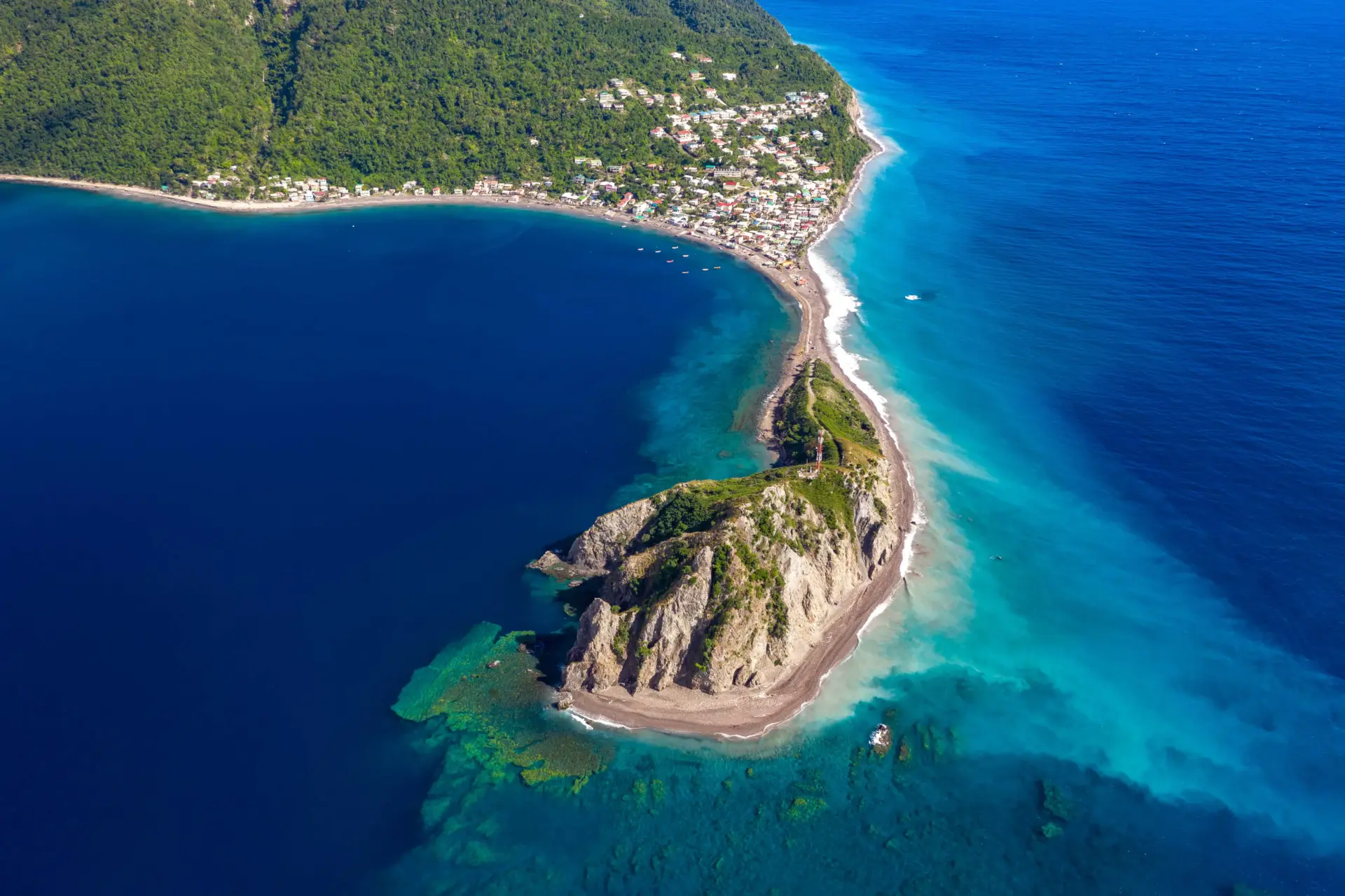 Most Stable CBI Programs: 2026 Guide 1 A striking bird's-eye view of a coastal landscape in Dominica. A thin, curved strip of beach and road connects a densely populated coastal village nestled against steep, jungle-covered mountains to a rocky, green headland in the foreground. The water to the left of the land bridge is a calm, dark blue, while the ocean to the right transitions into brilliant shades of light turquoise and aquamarine with visible coral reefs below the surface.