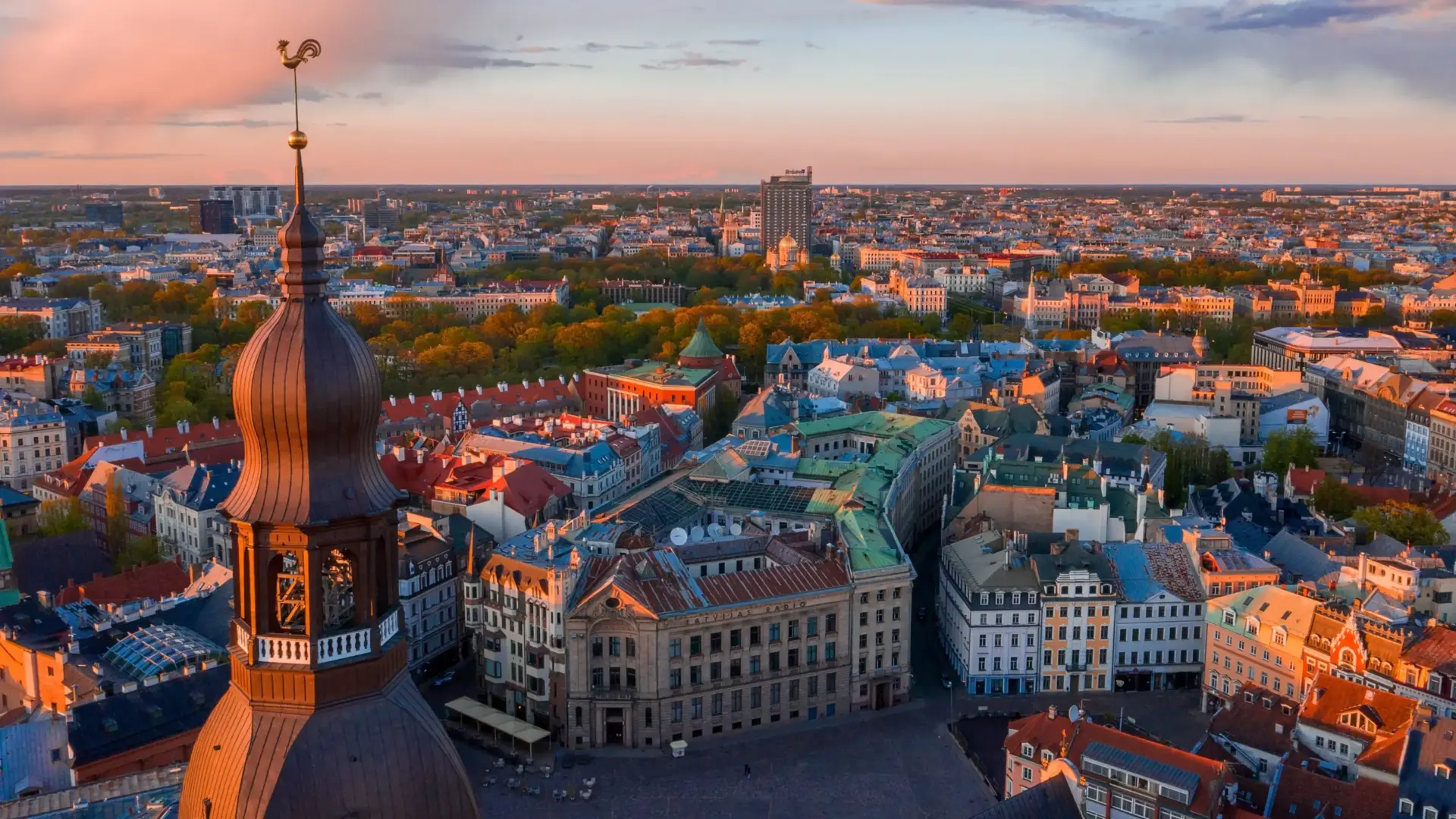 A beautiful sunset panoramic view of Riga, Latvia. The foreground is dominated on the left by the detailed, tiered wooden architecture of a church spire topped with a distinctive golden rooster. Below, the sprawling historic town square is lined with multi-story European buildings featuring red-orange roofs, teal-accented roofs, and classic facades catching the warm evening light. A dense band of lush green trees cuts through the middle ground, separating the old city from the modern skyline in the distance under a soft orange and pink sky.